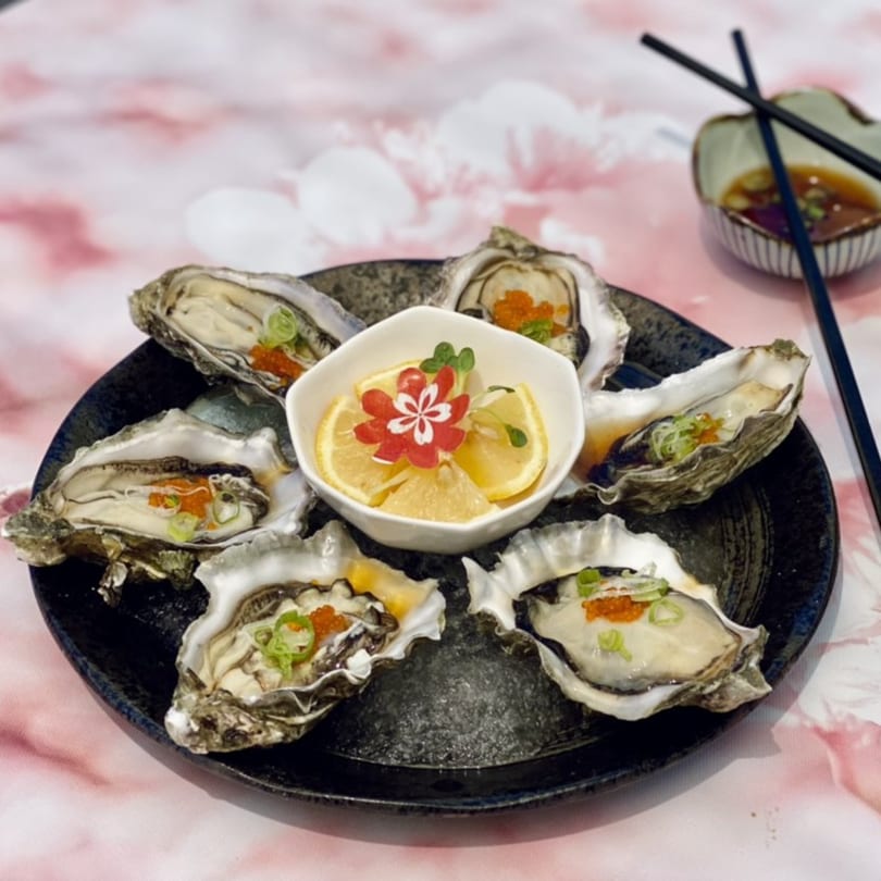 Six fresh oysters on a black plate with lemon slices, garnished with roe and green onions.