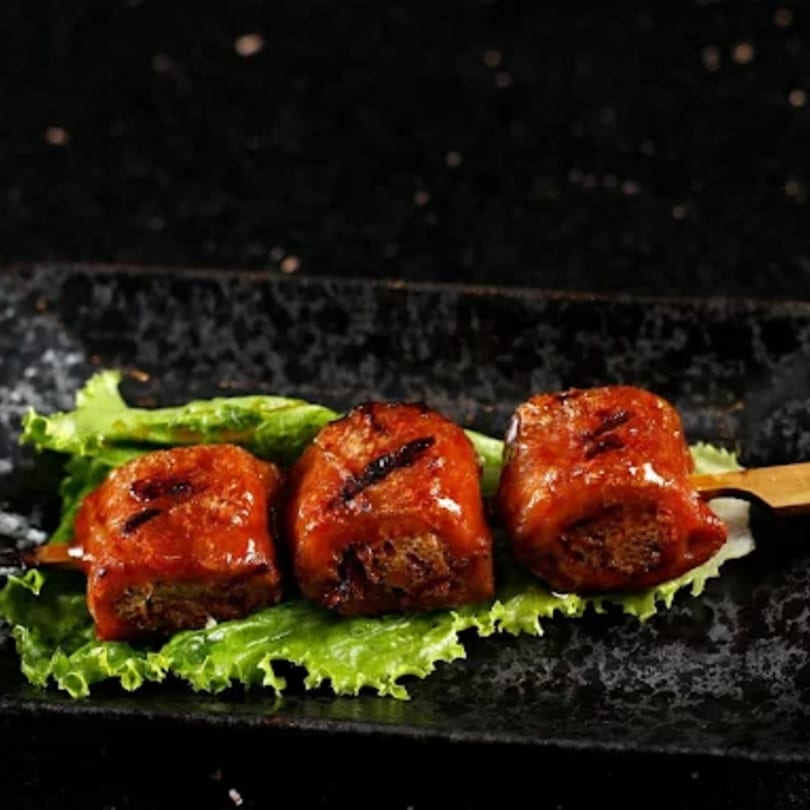 Three glazed Japanese skewers, possibly yakitori, on a bed of lettuce on a dark plate.